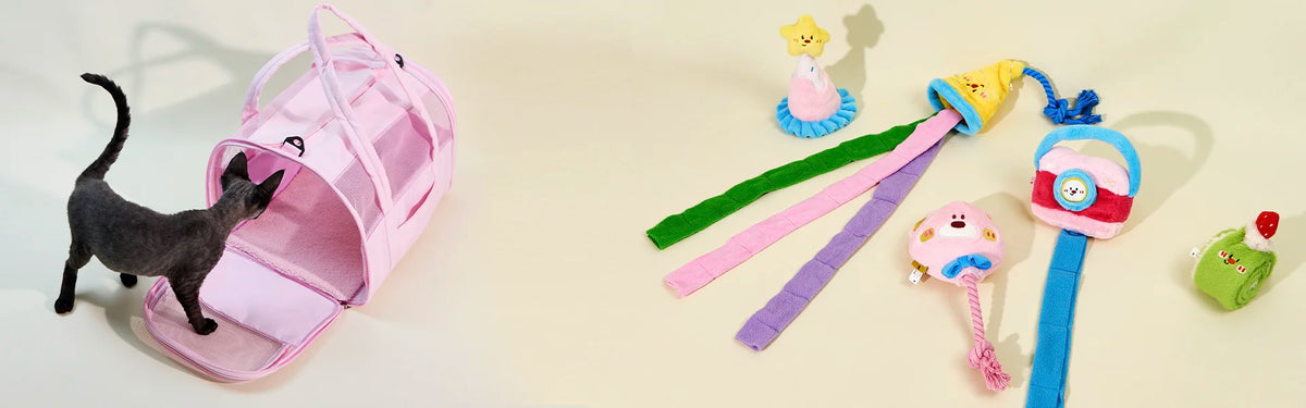 A black cat exploring an open pink mesh pet carrier bag next to colorful plush pet toys, including a birthday hat with long ribbons and a camera plush toy.