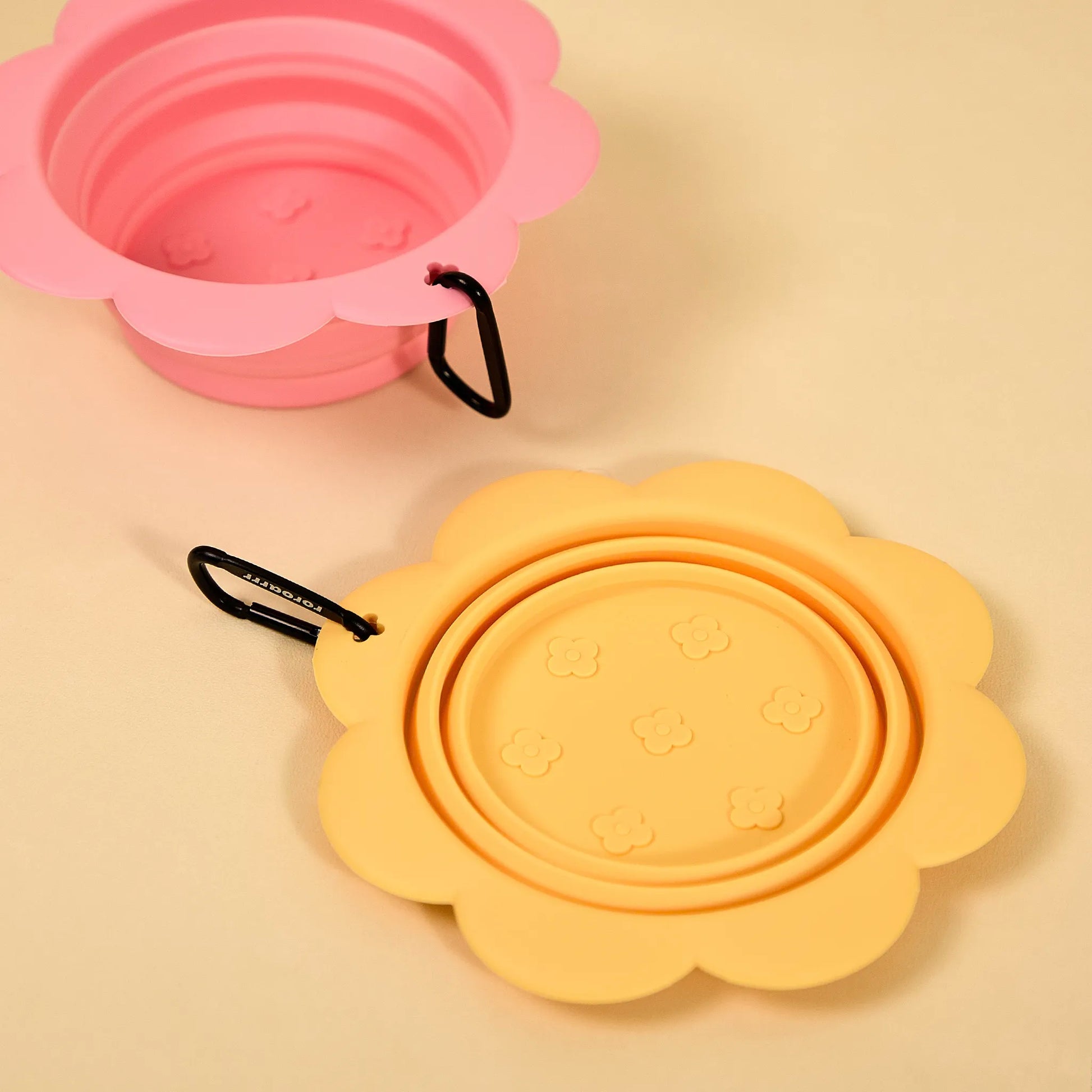 Pink and yellow collapsible flower-shaped silicone pet bowls shown open and folded on a tan background